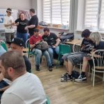 Uczestnicy turnieju Pro Evolution Soccer 6 organizowanego przez Brzeską Scenę Gier Komputerowych (BSGK) grają na konsolach w sali turniejowej w Brzegu. Gracze siedzą przy stołach z kontrolerami w dłoniach, w tle widoczny baner BSGK oraz inni uczestnicy wydarzenia.