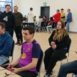 Turniej FC 26 w Skarbimierzu – uczestnicy siedzą przy stołach i grają na konsolach, podczas gdy inni stoją i obserwują rozgrywki w jasnej sali z dużymi oknami.